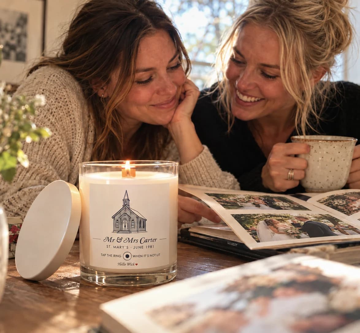 Two grown daughters at a sunlit table over their parents' wedding album, a lit Hello Wick candle labelled Mr & Mrs Carter beside them