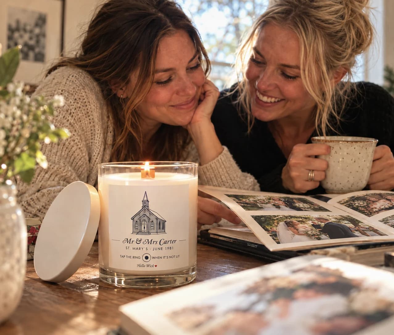 Two grown daughters at a sunlit table over their parents' wedding album, a lit Hello Wick candle labelled Mr & Mrs Carter beside them