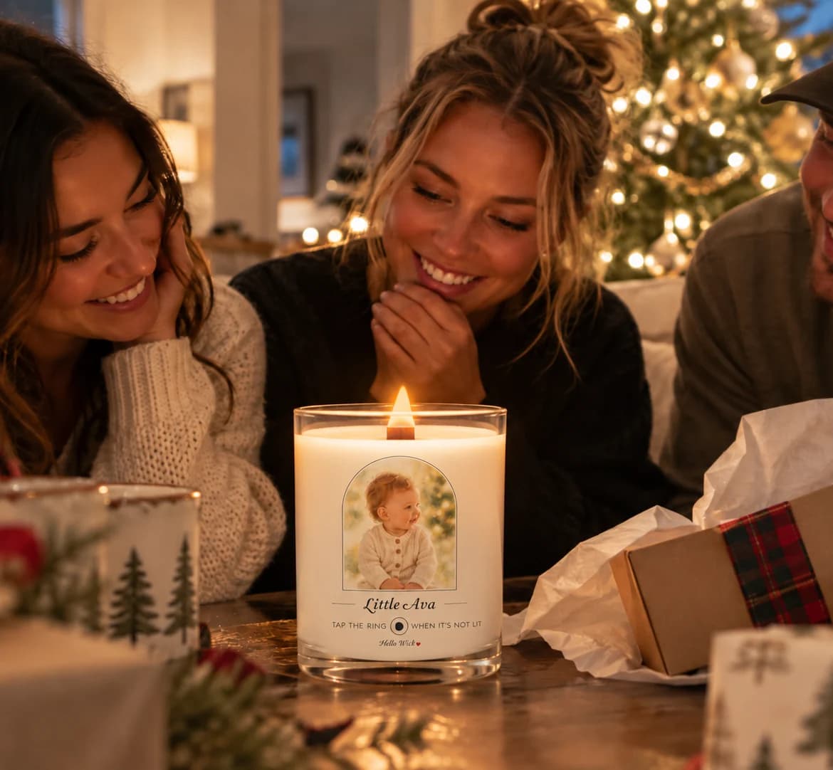 Three young adults at a candlelit coffee table by the Christmas tree, smiling at a lit Hello Wick candle labelled Little Ava with the baby’s photo in the arch, a gift box open beside it