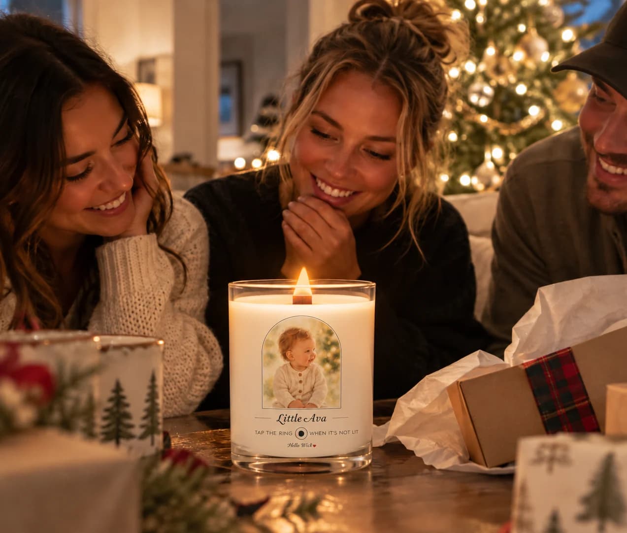 Three young adults at a candlelit coffee table by the Christmas tree, smiling at a lit Hello Wick candle labelled Little Ava with the baby’s photo in the arch, a gift box open beside it