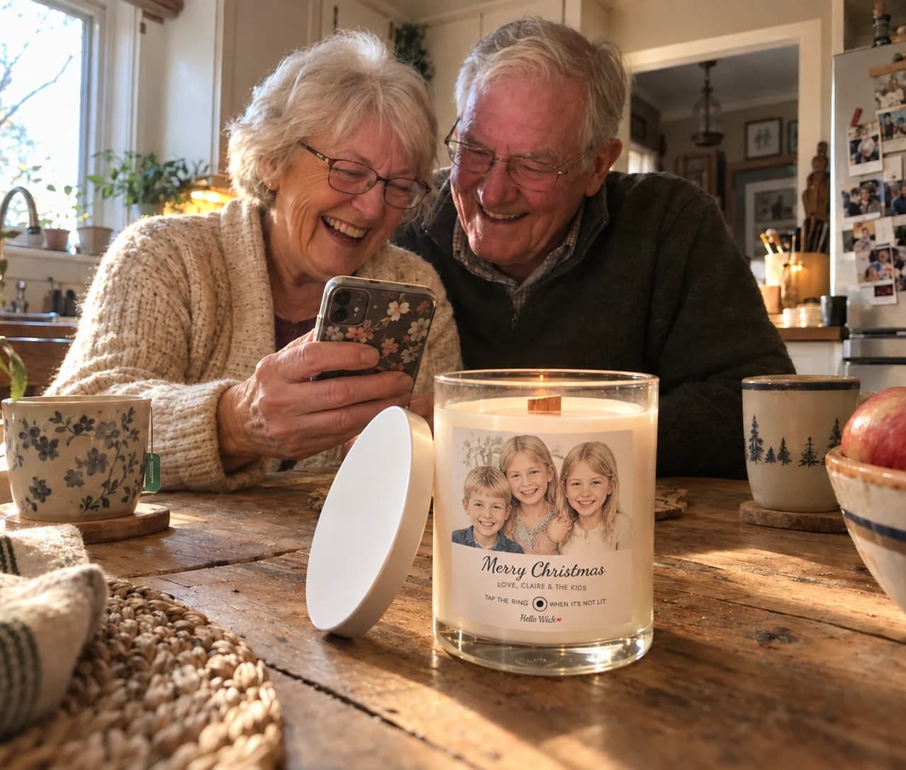 Grandparents at their kitchen table laughing at a phone, a lit Hello Wick candle labeled Merry Christmas from the grandkids beside them